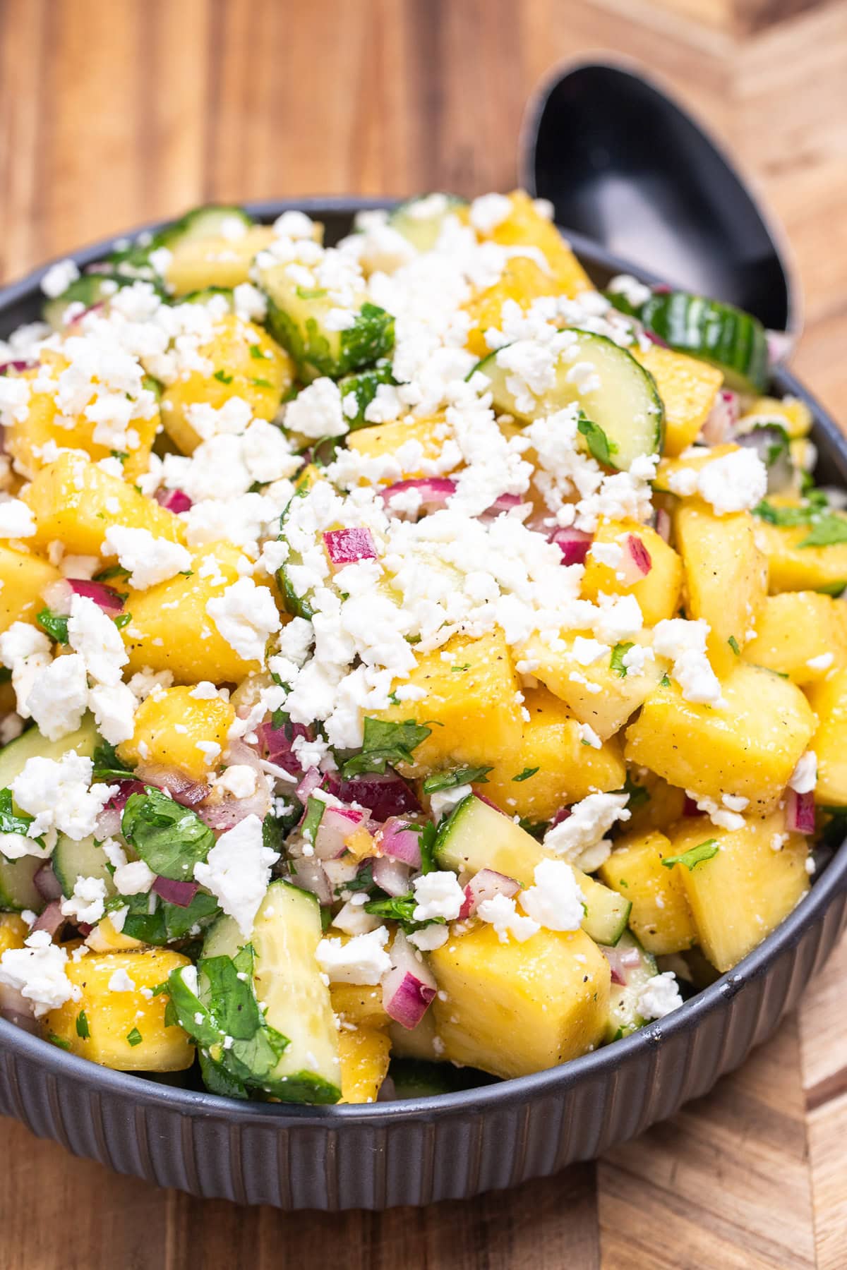 A bowl of pineapple cucumber salad topped with feta cheese sitting on a wooden cutting board with a spoon behind it.