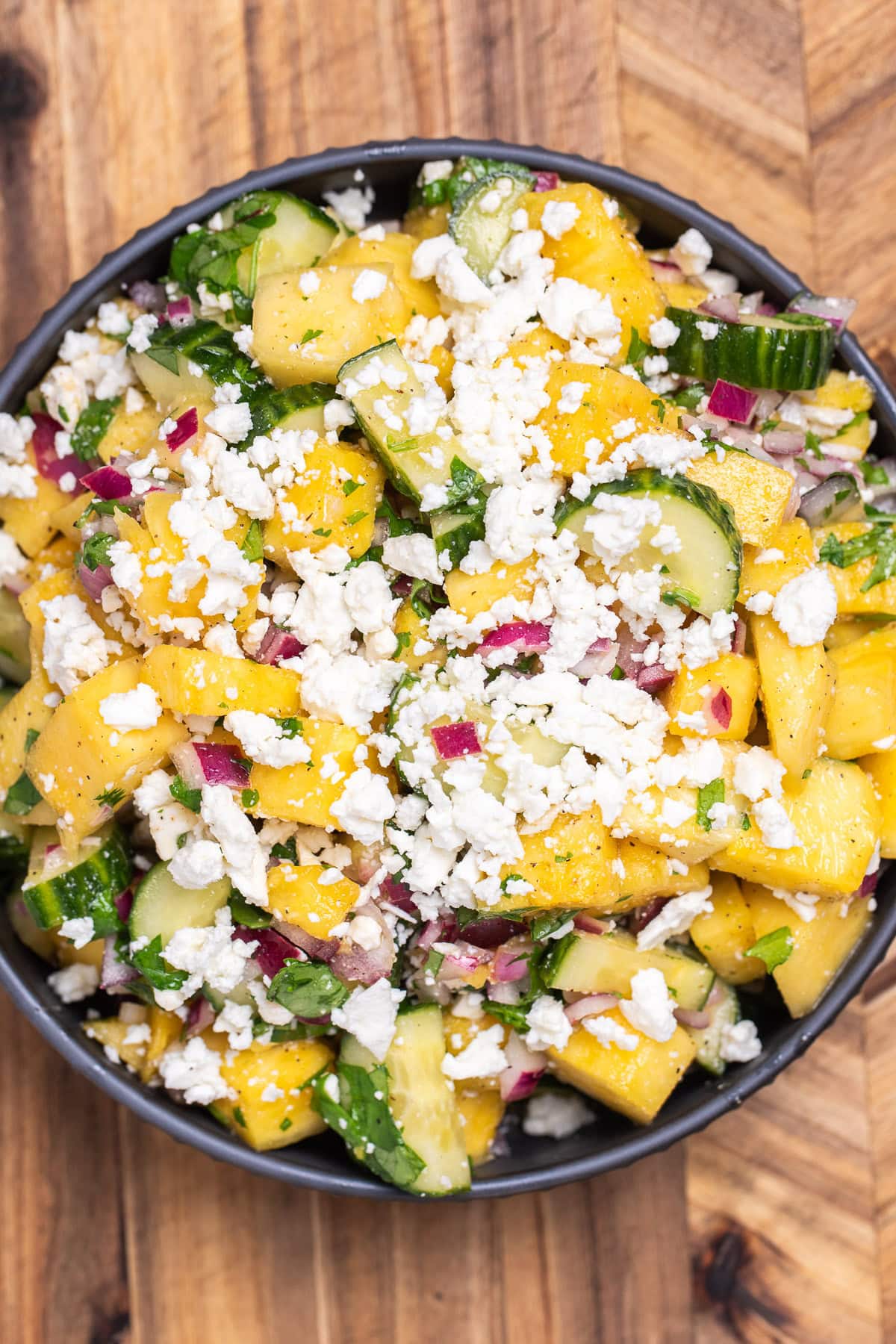 A bowl of pineapple cucumber salad topped with feta, sitting on a wooden cutting board.