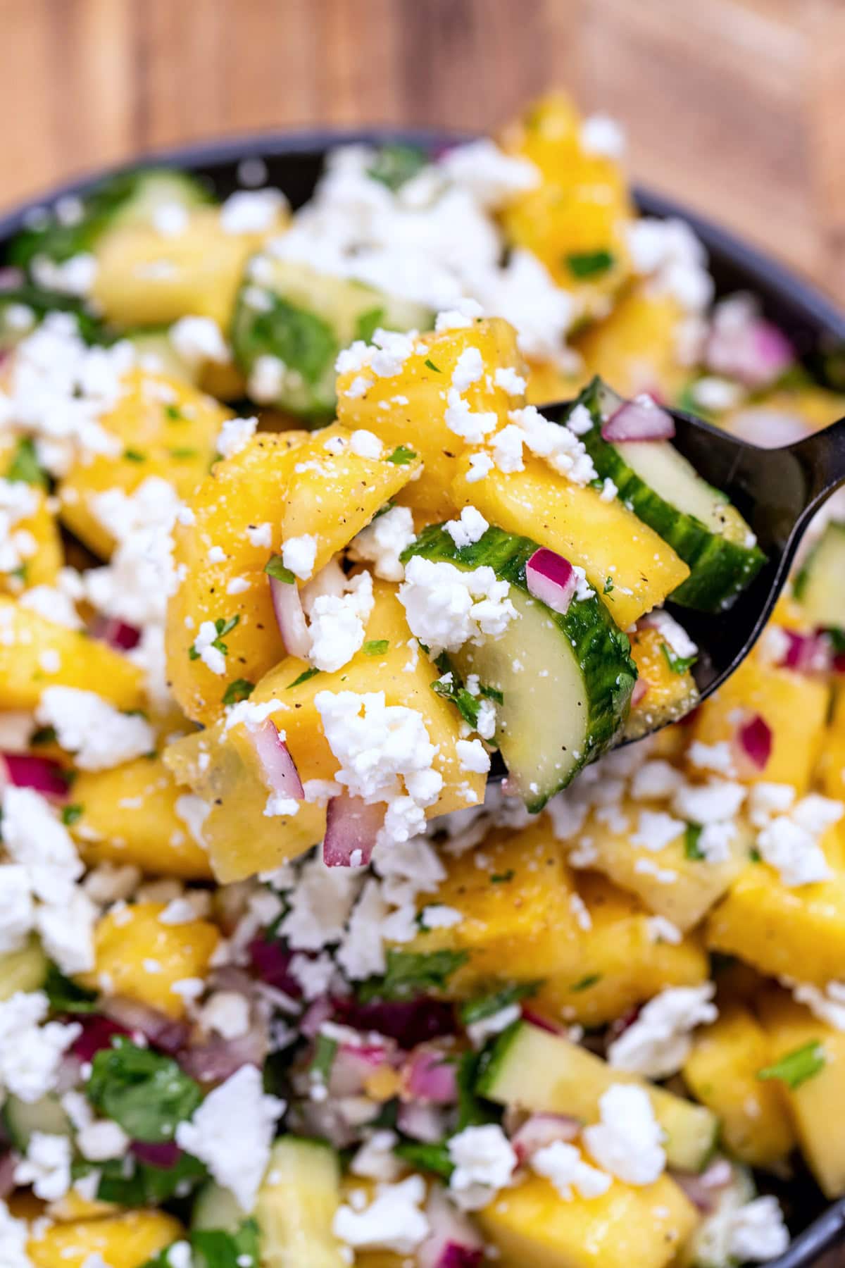 A bowl of pineapple cucumber salad topped with feta cheese with a spoon lifting some up.
