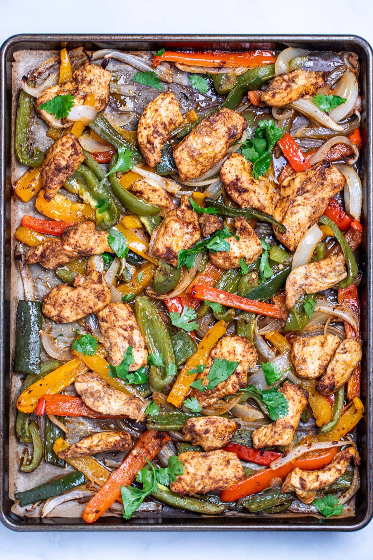 A sheet pan lined with parchment paper with cooked chicken and peppers topped with fresh cilantro.