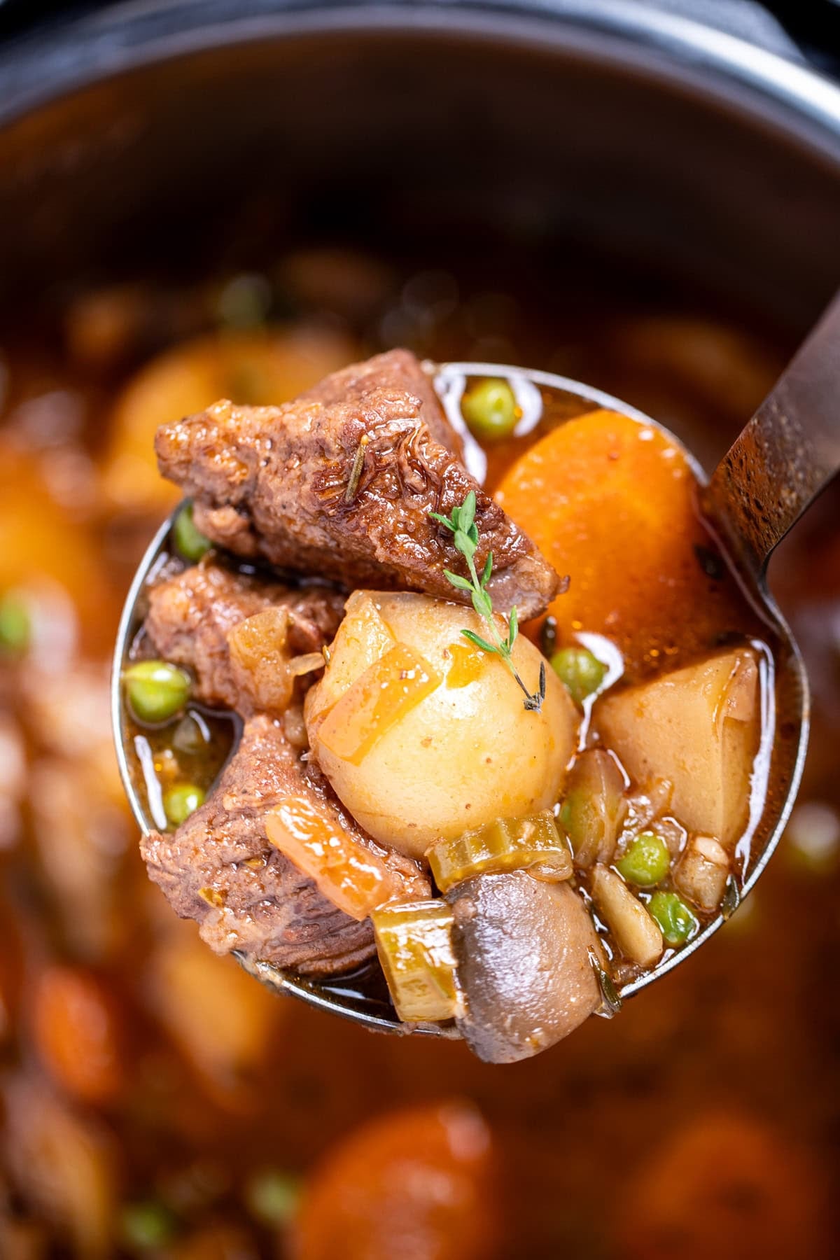 A ladle lifting up beef stew with beef, potatoes, carrots, peas, and mushrooms, topped with fresh thyme.