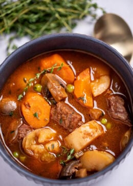A bowl of beef stew with potatoes, carrots, peas, beef, and mushrooms in a tomato based broth topped with fresh thyme, with a spoon and fresh herbs in the background.