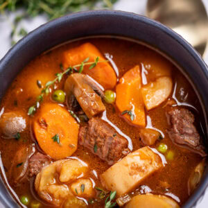 A bowl of beef stew with potatoes, carrots, peas, beef, and mushrooms in a tomato based broth topped with fresh thyme, with a spoon and fresh herbs in the background.