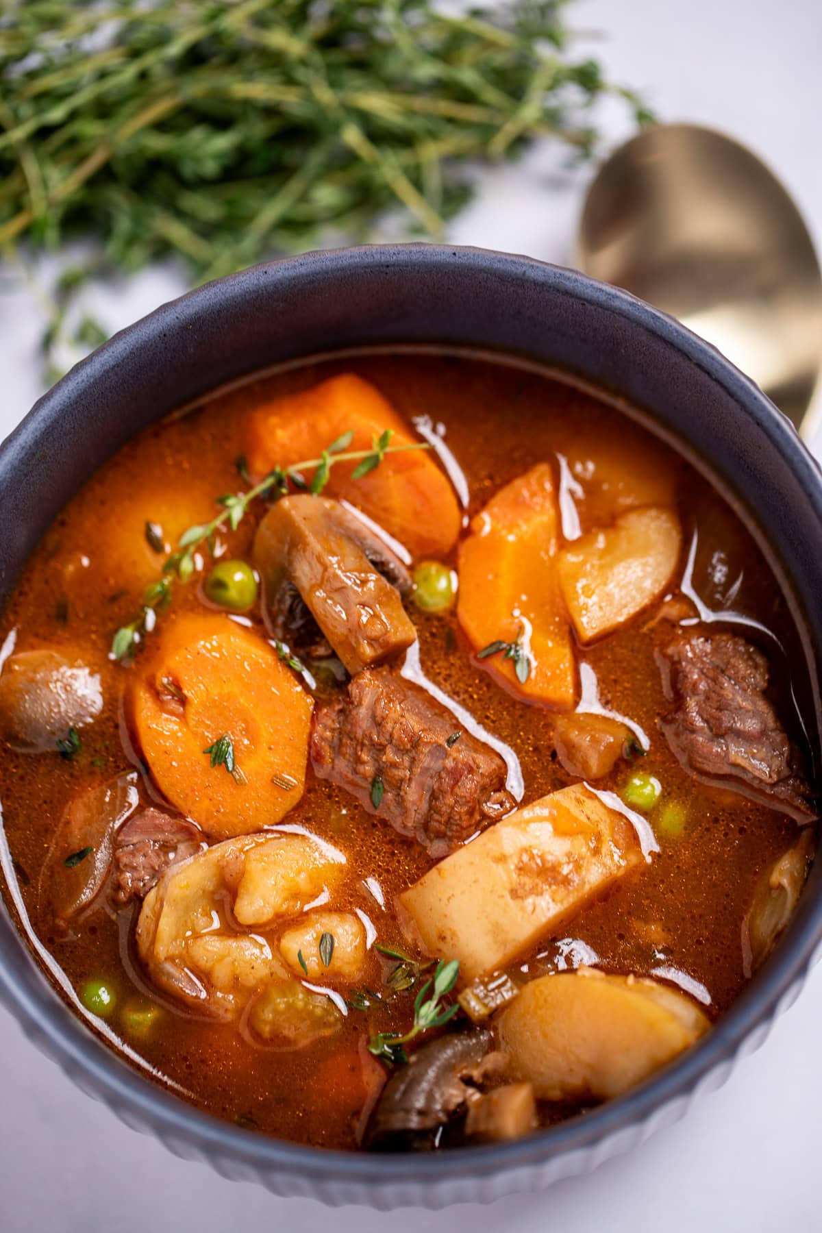 A bowl of beef stew with potatoes, carrots, peas, beef, and mushrooms in a tomato based broth topped with fresh thyme, with a spoon and fresh herbs in the background.