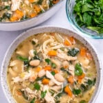 A bowl of white bean chicken soup with a pot of soup in the top left of the photo, and a small glass bowl of chopped parsley on the top right.