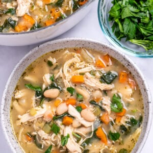 A bowl of white bean chicken soup with a pot of soup in the top left of the photo, and a small glass bowl of chopped parsley on the top right.