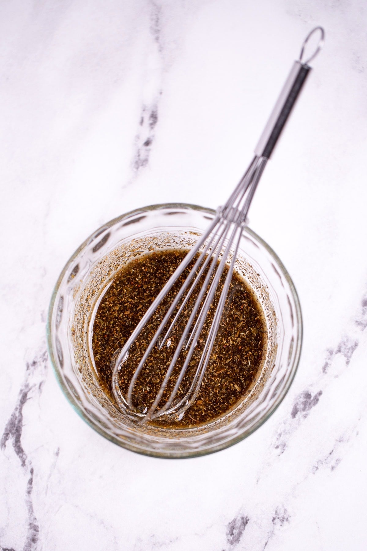 A small glass bowl on a table with olive oil and spices whisked together, with a whisk resting in the bowl.