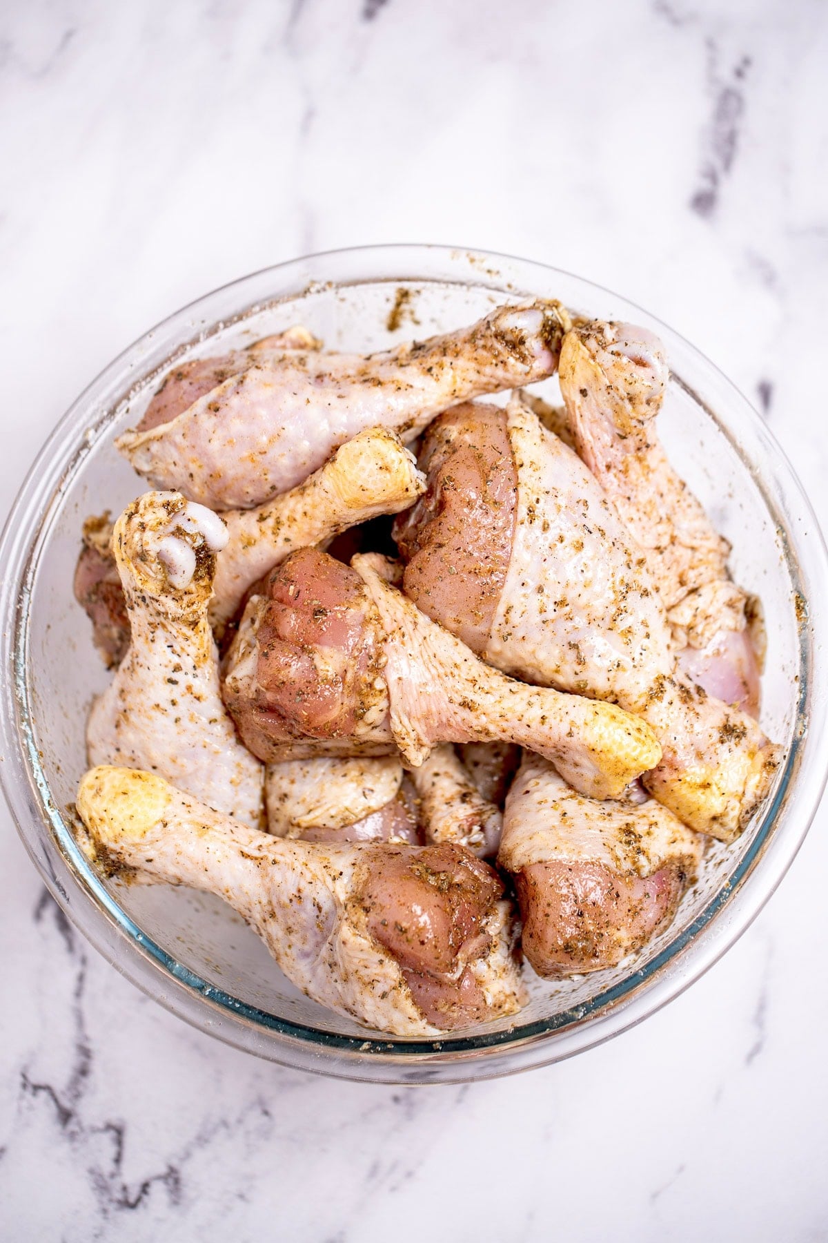 A glass bowl on a table, full of raw chicken drumsticks coated in olive oil and seasoning.