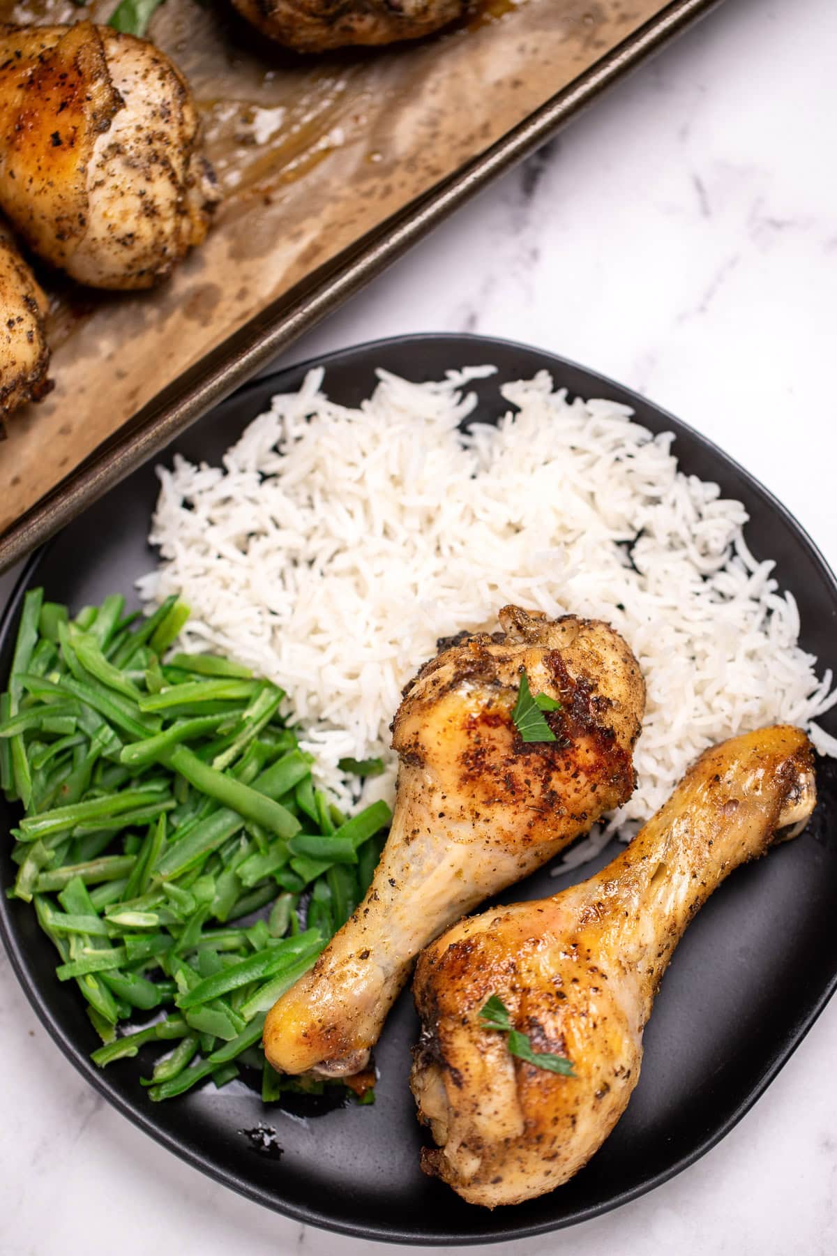 Two baked chicken legs on a plate with rice and green beans, in front of a sheet pan with more chicken drumsticks.