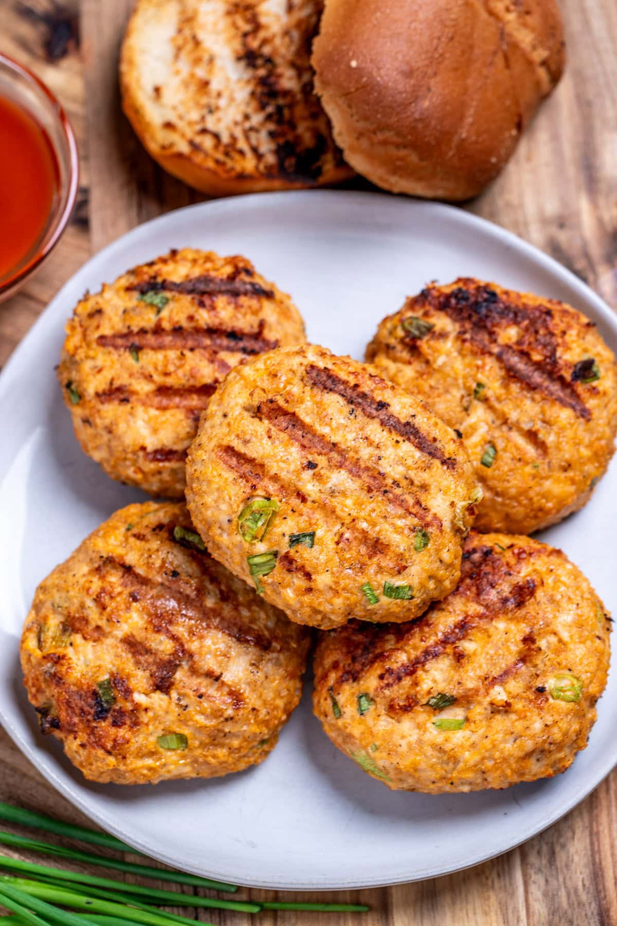 A plate of buffalo chicken burger patties on a cutting board next to a bowl of buffalo sauce and chives.