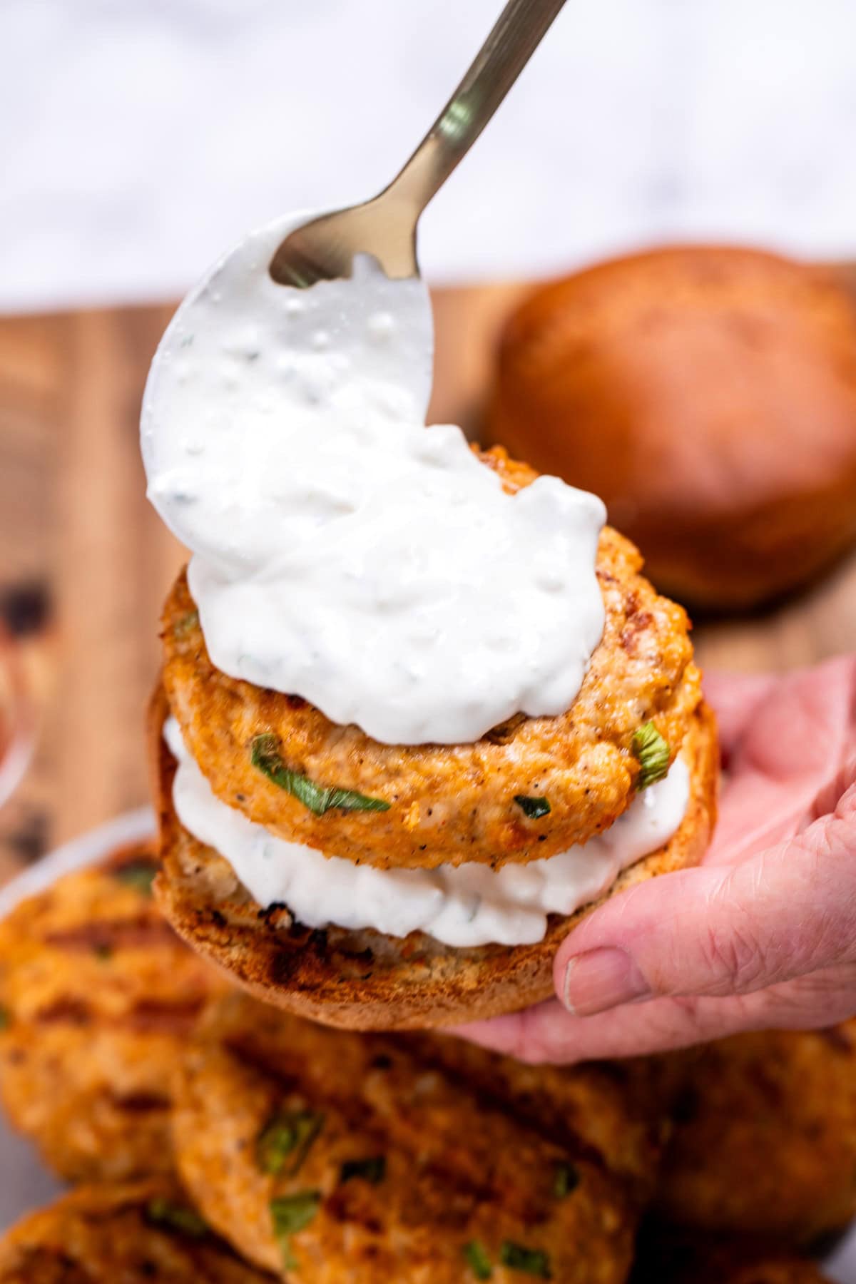 A hand holding a buffalo chicken burger with a spoon placing blue cheese dressing on the burger, over a plate of burger patties.