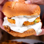 A buffalo chicken burger on a bun with blue cheese dripping from the burger, and two hands holding the burger from the back.