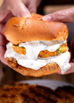 A buffalo chicken burger on a bun with blue cheese dripping from the burger, and two hands holding the burger from the back.