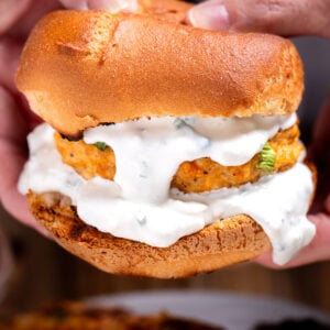 A buffalo chicken burger on a bun with blue cheese dripping from the burger, and two hands holding the burger from the back.