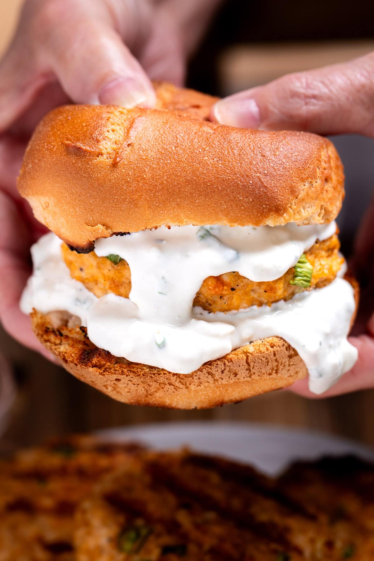 A buffalo chicken burger on a bun with blue cheese dripping from the burger, and two hands holding the burger from the back.