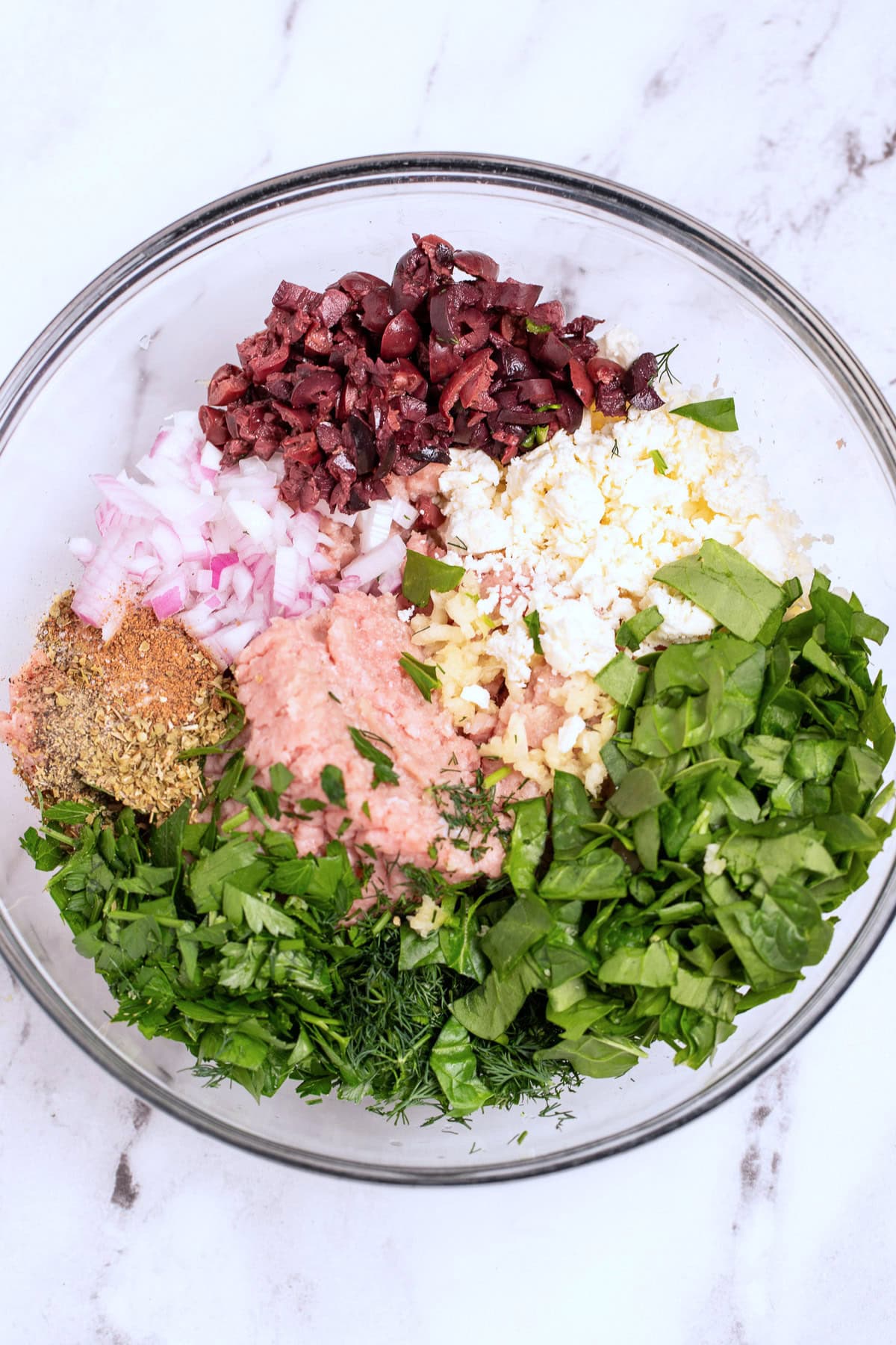 A glass bowl on a table with all of the greek turkey burger ingredients before being mixed together.