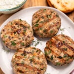 A plate resting on a cutting board, with four grilled greek turkey burgers, next to red onion, a burger bun, and a bowl of tzatziki on the board.