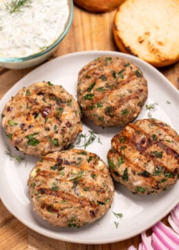 A plate resting on a cutting board, with four grilled greek turkey burgers, next to red onion, a burger bun, and a bowl of tzatziki on the board.