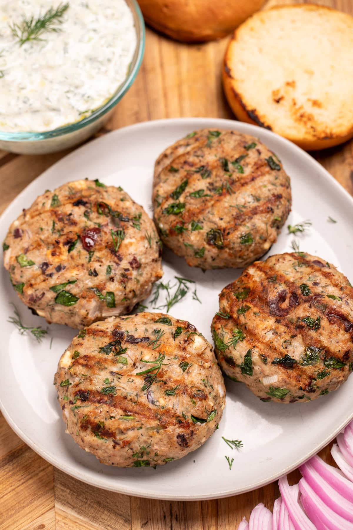 A plate resting on a cutting board, with four grilled greek turkey burgers, next to red onion, a burger bun, and a bowl of tzatziki on the board.