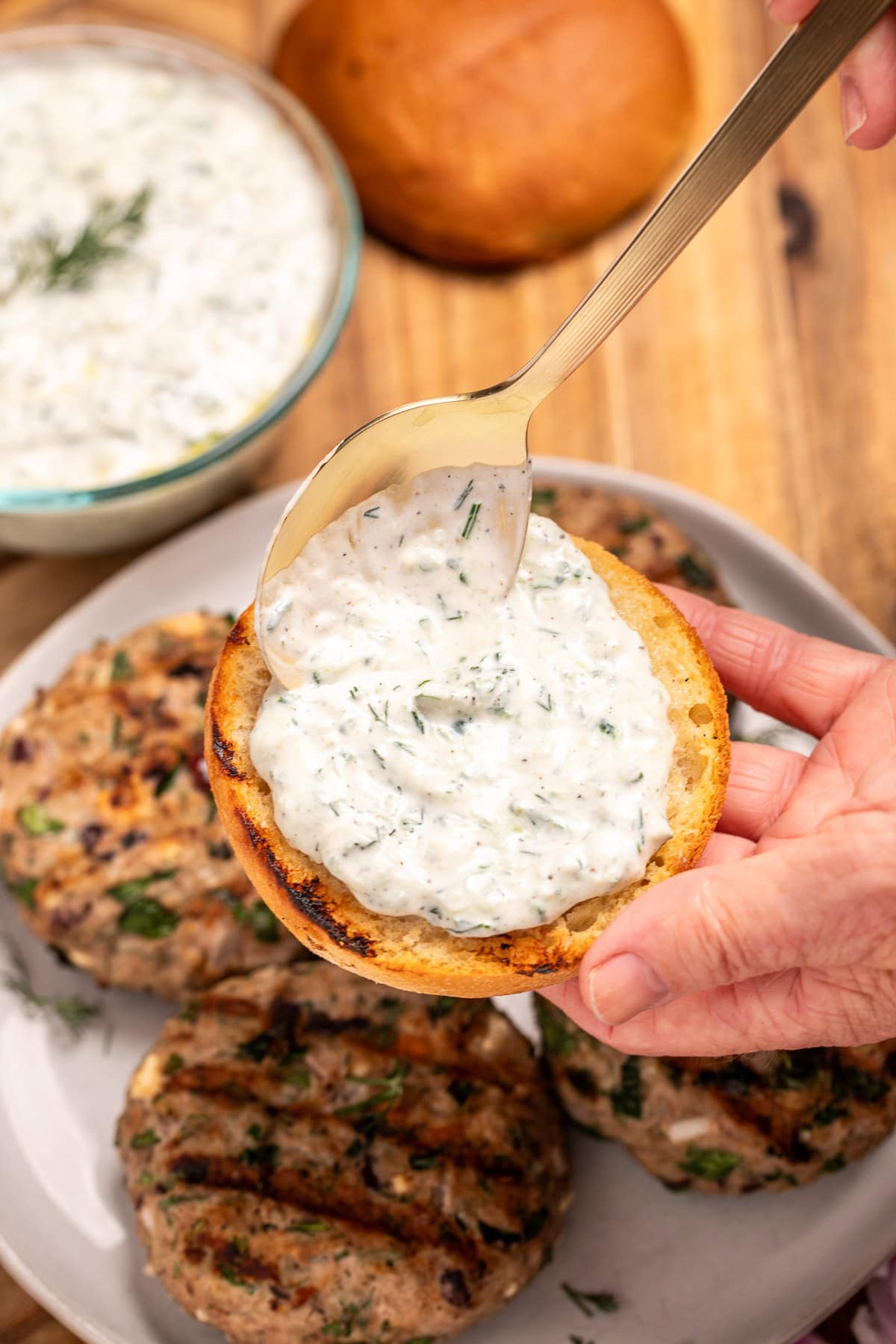 Hands spooning tzatziki sauce onto a burger bun above a plate resting on a cutting board, with four grilled greek turkey burgers, next to red onion, a burger bun, and a bowl of tzatziki on the board.