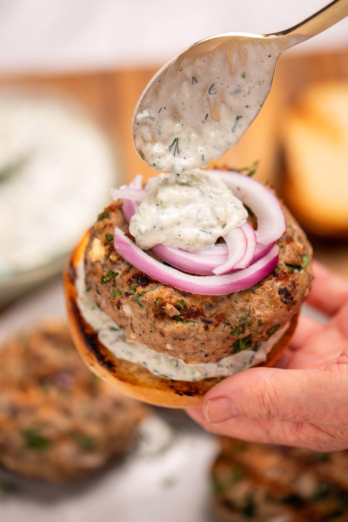 A hand spooning tzatziki sauce onto a greek turkey burger with red onion, above a plate of burger patties.