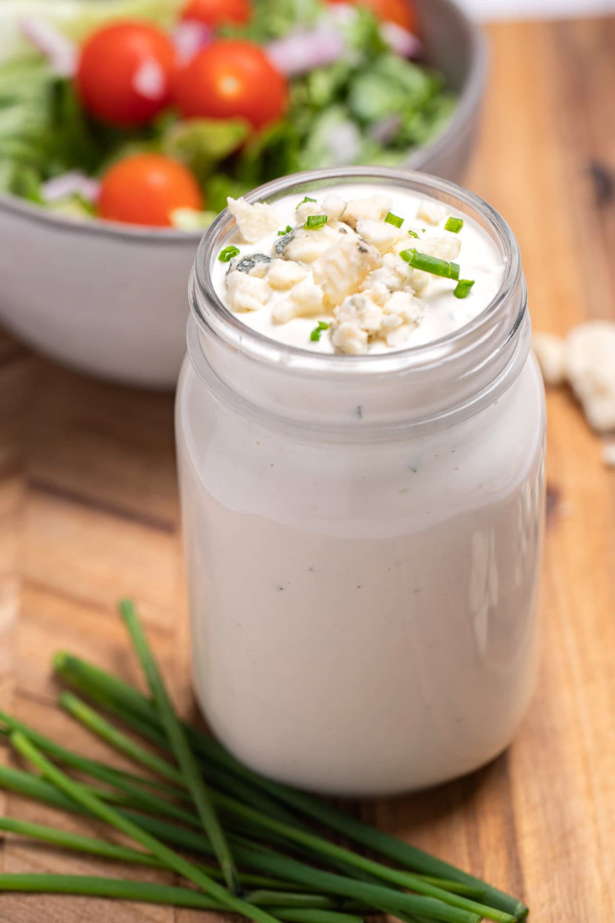 A mason jar of homemade blue cheese dressing resting on a cutting board with fresh chives and blue cheese crumbles around it, and a bowl of salad in the background.