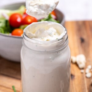 A mason jar of homemade blue cheese dressing resting on a cutting board with fresh chives and blue cheese crumbles around it, and a bowl of salad in the background. A spoon is lifting some of the dressing out of the jar.