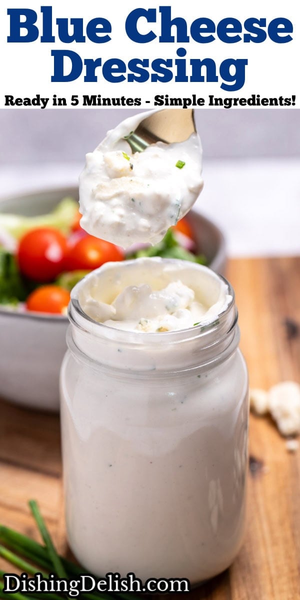 Pinterest pin with a mason jar of homemade blue cheese dressing resting on a cutting board with fresh chives and blue cheese crumbles around it, and a bowl of salad in the background. A spoon is lifting some of the dressing out of the jar.