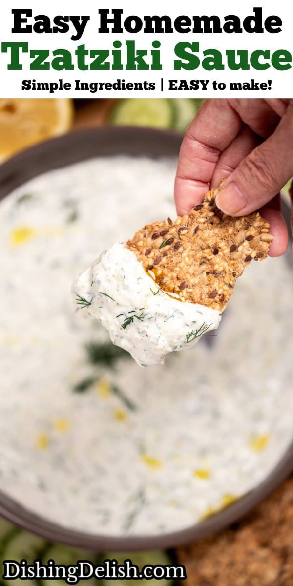 Pinterest pin with a bowl of homemade tzatziki sauce topped with fresh dill, on a cutting board with sliced cucumber, lemon, and gluten free crackers, with a hand holding a cracker lifting some tzatziki up.