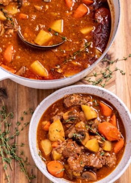 A bowl of Irish stew on a cutting board, topped with fresh thyme, and a pot of stew behind it.