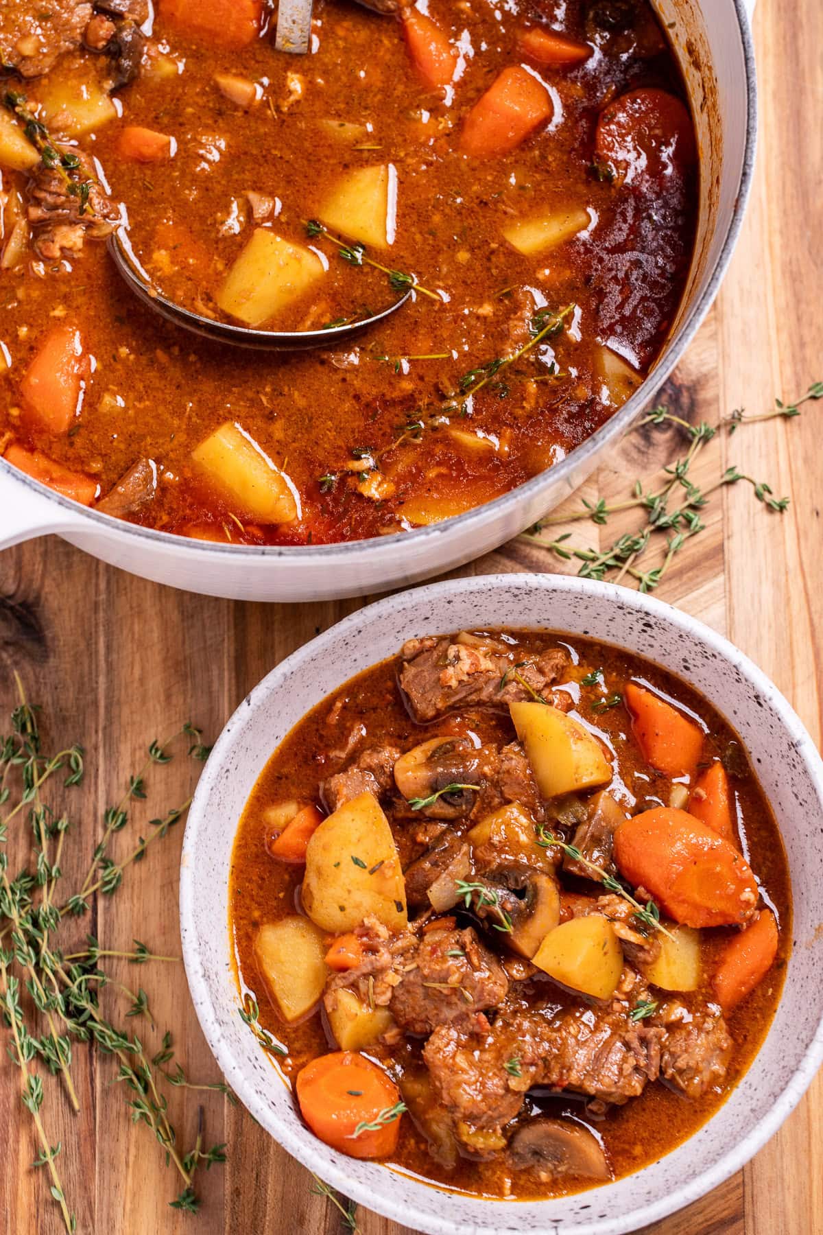 A bowl of Irish stew on a cutting board, topped with fresh thyme, and a pot of stew behind it.