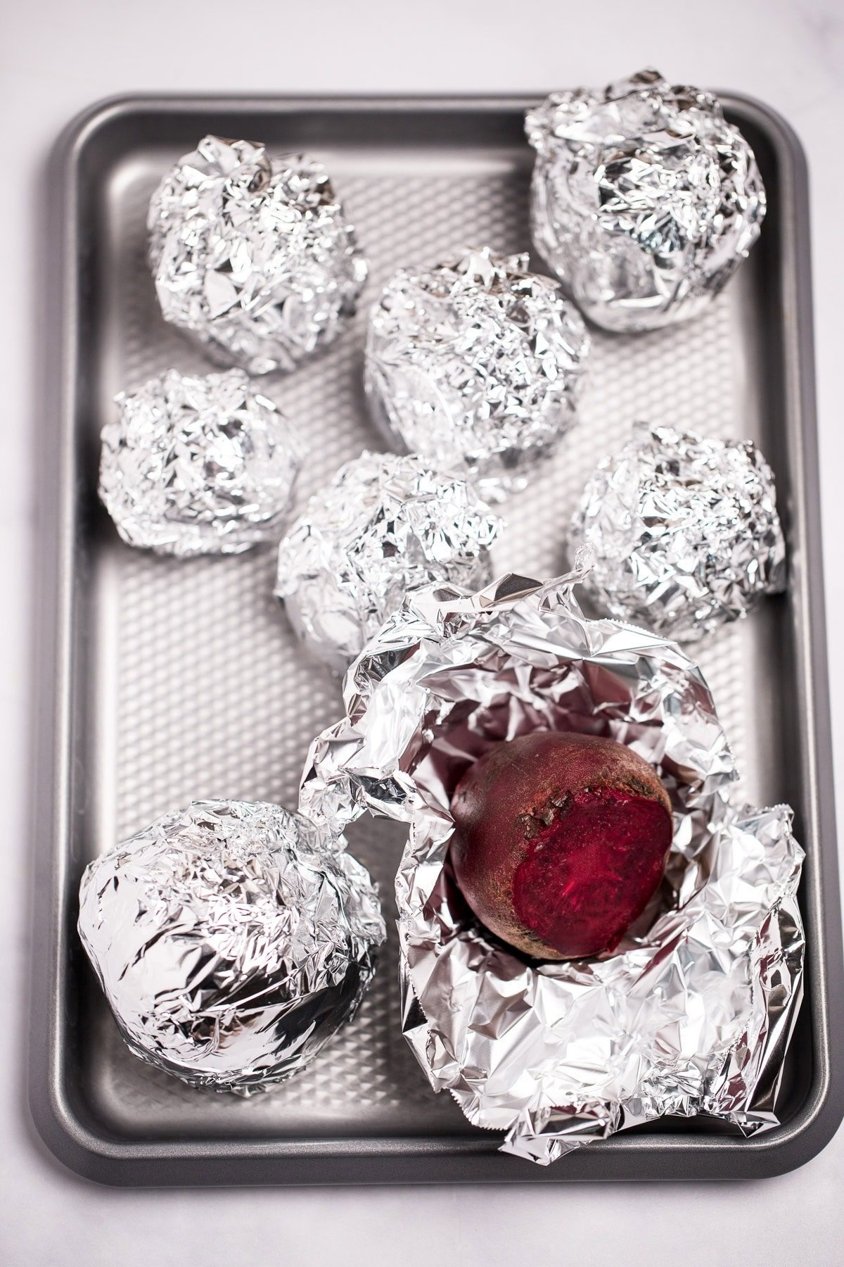 A baking sheet with beets wrapped in tinfoil, with one open to show the beet, before baking.