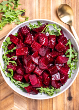 A bowl of roasted beet salad resting on arugula, on a cutting board next to a spoon.