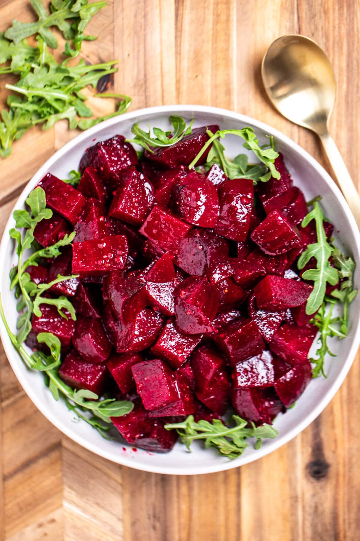A bowl of roasted beet salad resting on arugula, on a cutting board next to a spoon.