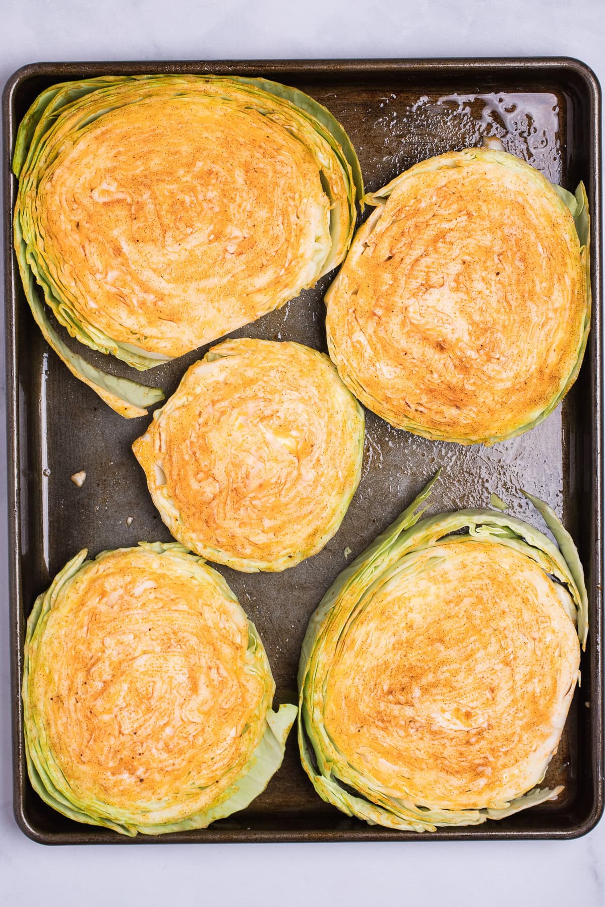 A sheet pan of raw cabbage steaks topped with olive oil and seasoning before being roasted.