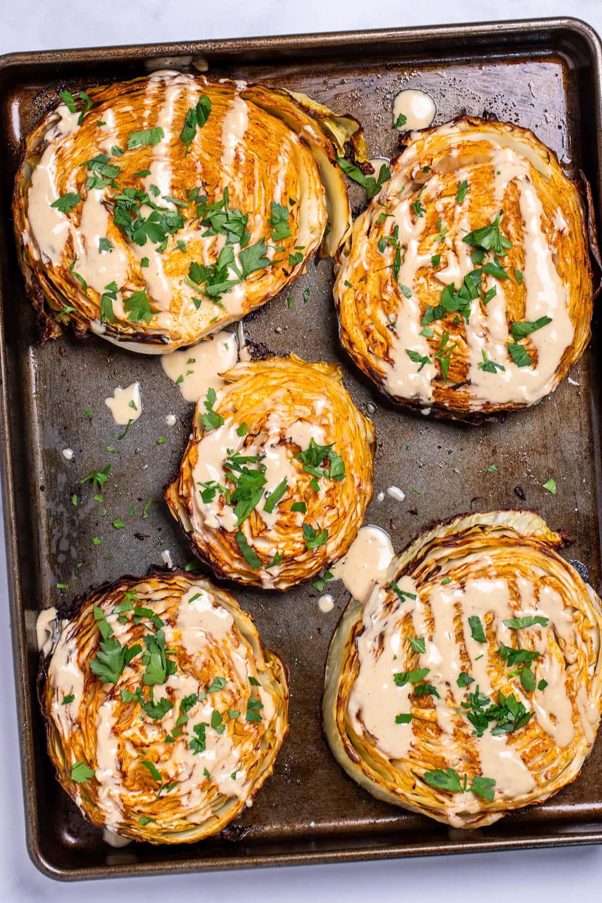 A sheet pan of roasted cabbage steaks topped with tahini dressing and chopped fresh parsley.
