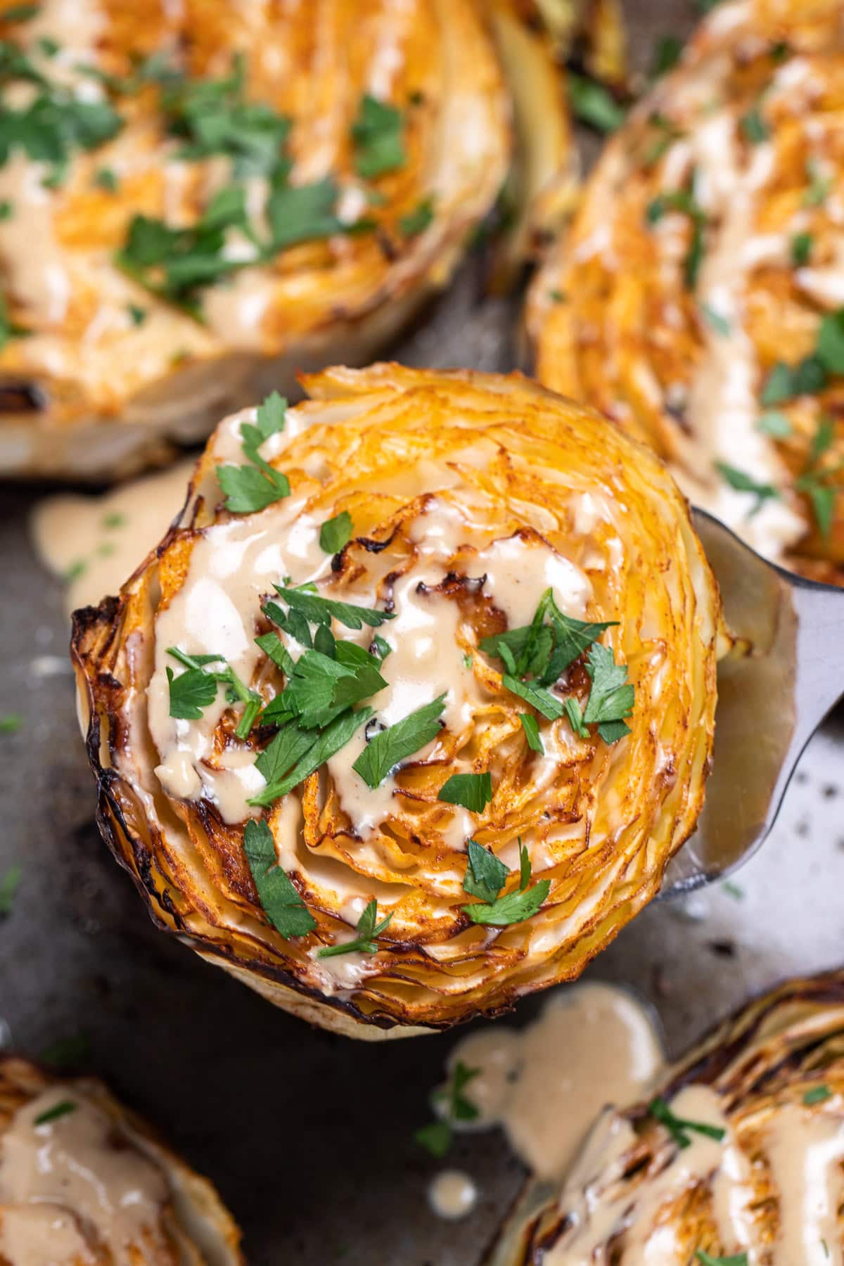 A roasted cabbage steak topped with tahini dressing and chopped fresh parsley, over a sheet pan of cabbage steaks.