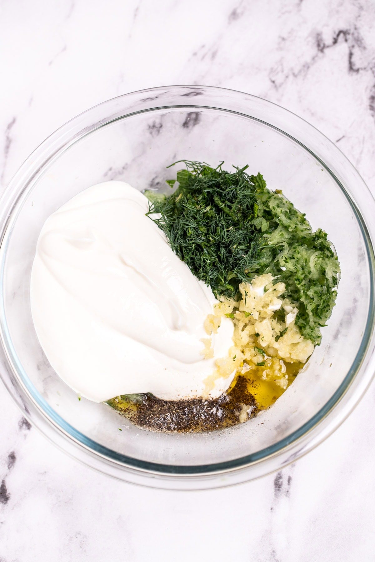 Glass bowl on a table with individual ingredients for tzatziki sauce before being mixed together.