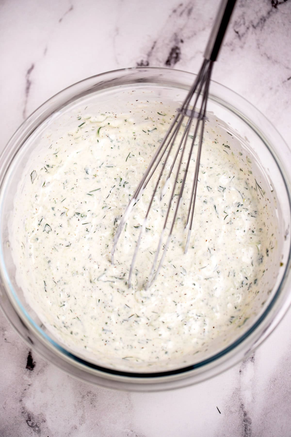 Glass bowl on a table with tzatziki dressing and a whisk resting in it.