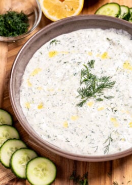 A bowl of homemade tzatziki sauce topped with fresh dill, on a cutting board with sliced cucumber, lemon, and a small bowl of chopped fresh dill.