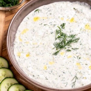 A bowl of homemade tzatziki sauce topped with fresh dill, on a cutting board with sliced cucumber, lemon, and a small bowl of chopped fresh dill.