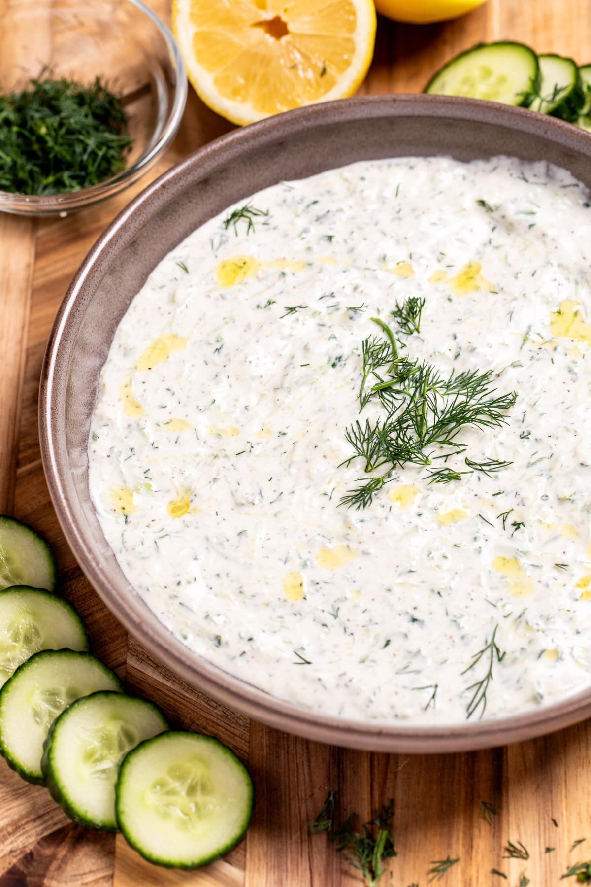 A bowl of homemade tzatziki sauce topped with fresh dill, on a cutting board with sliced cucumber, lemon, and a small bowl of chopped fresh dill.