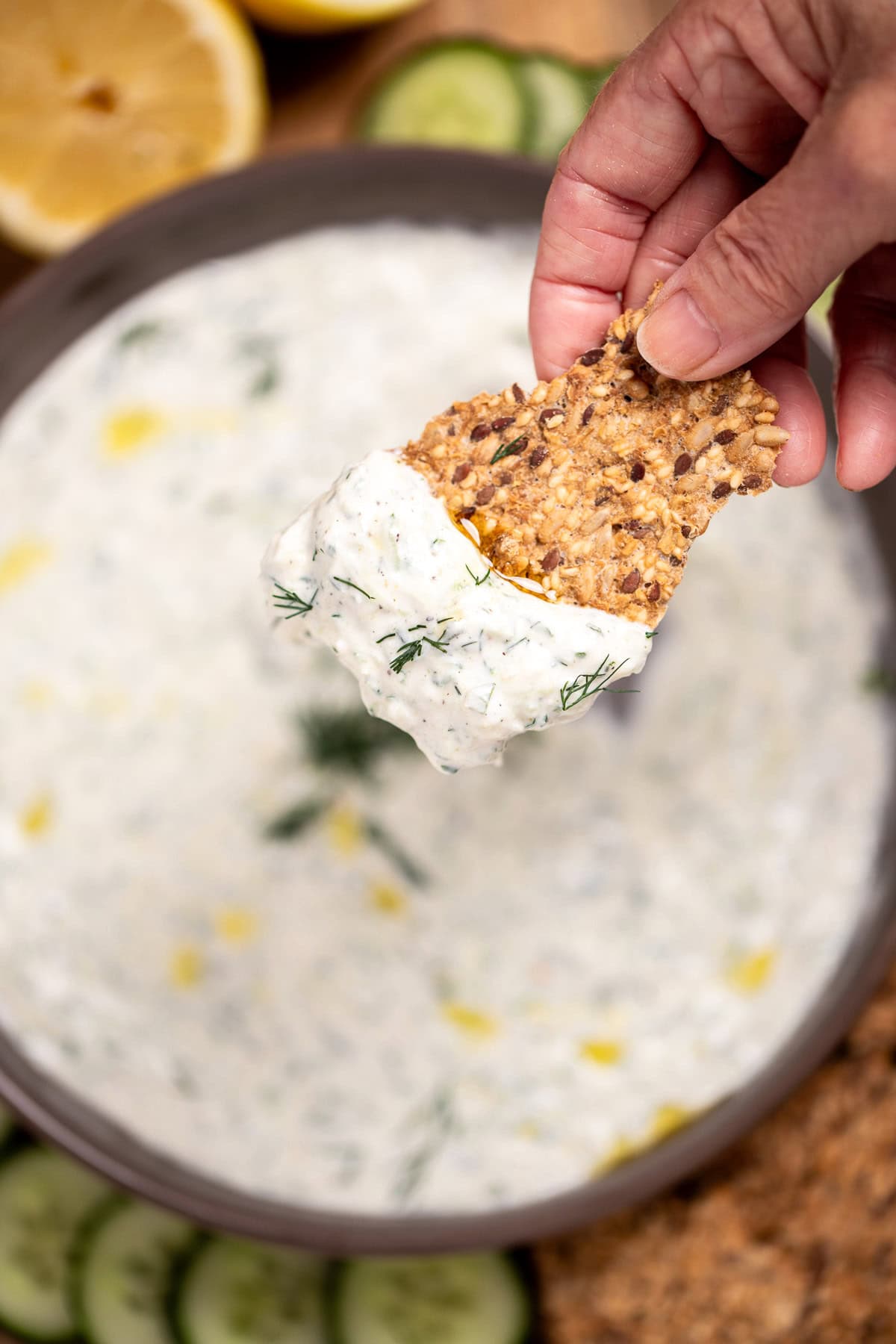 A bowl of homemade tzatziki sauce topped with fresh dill, on a cutting board with sliced cucumber, lemon, and gluten free crackers, with a hand holding a cracker lifting some tzatziki up.