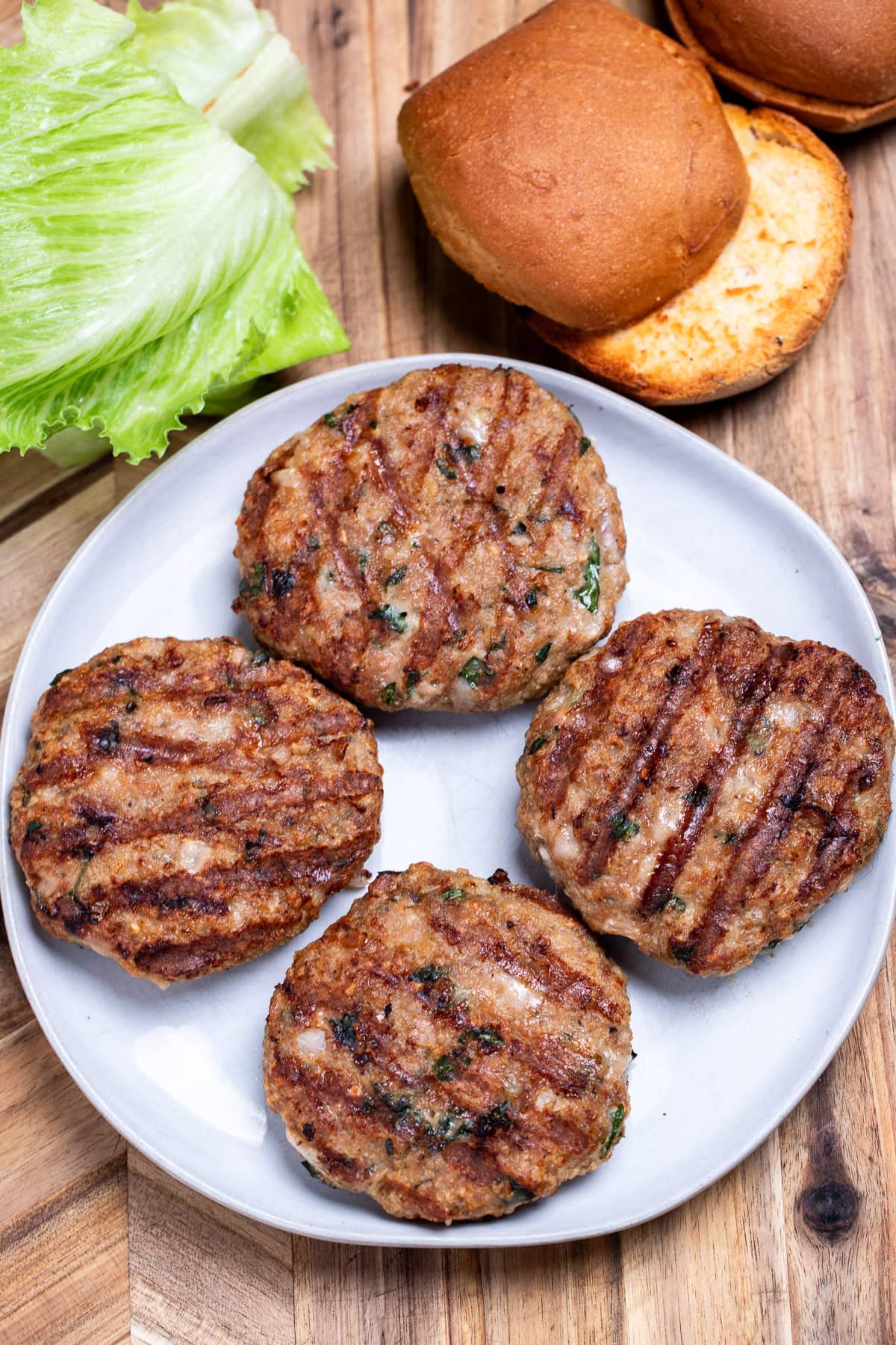 Four turkey burgers with grill marks on a plate resting on a wooden cutting board, next to lettuce and a hamburger bun.