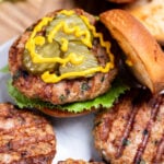 Four turkey burgers with grill marks on a plate, one in a burger bun with lettuce, pickles, and mustard, resting on a wooden cutting board, next to lettuce and a hamburger bun.