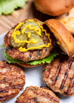Four turkey burgers with grill marks on a plate, one in a burger bun with lettuce, pickles, and mustard, resting on a wooden cutting board, next to lettuce and a hamburger bun.