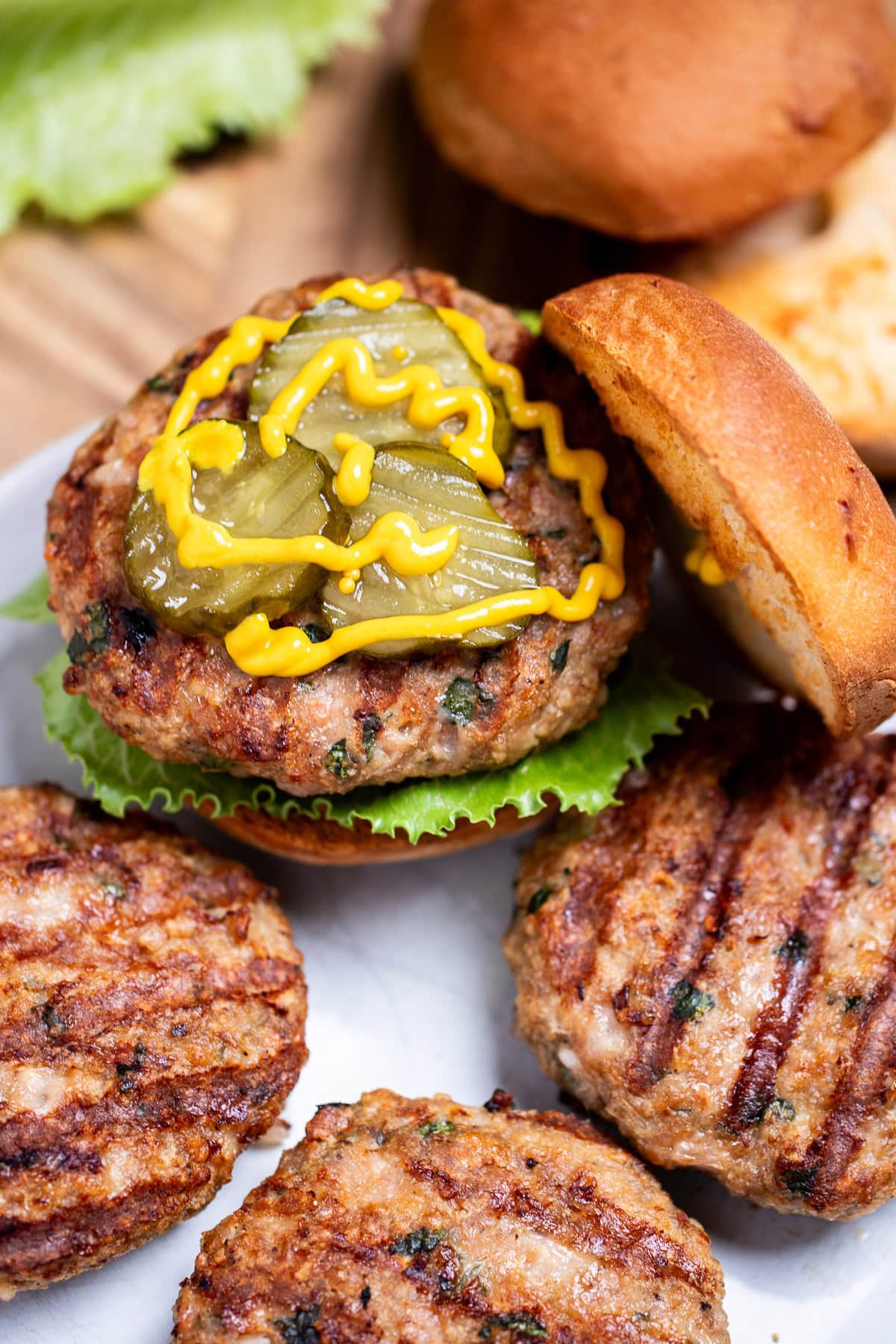 Four turkey burgers with grill marks on a plate, one in a burger bun with lettuce, pickles, and mustard, resting on a wooden cutting board, next to lettuce and a hamburger bun.