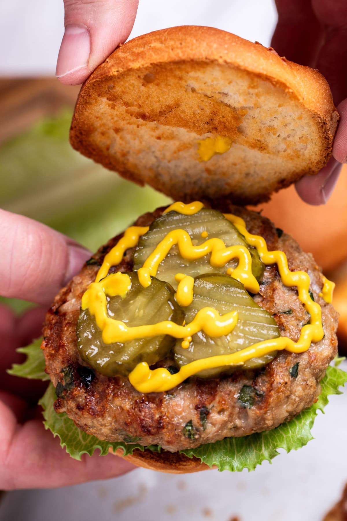 Two hands holding a grilled turkey burger with lettuce, pickles, and mustard, lifting the top bun piece up.