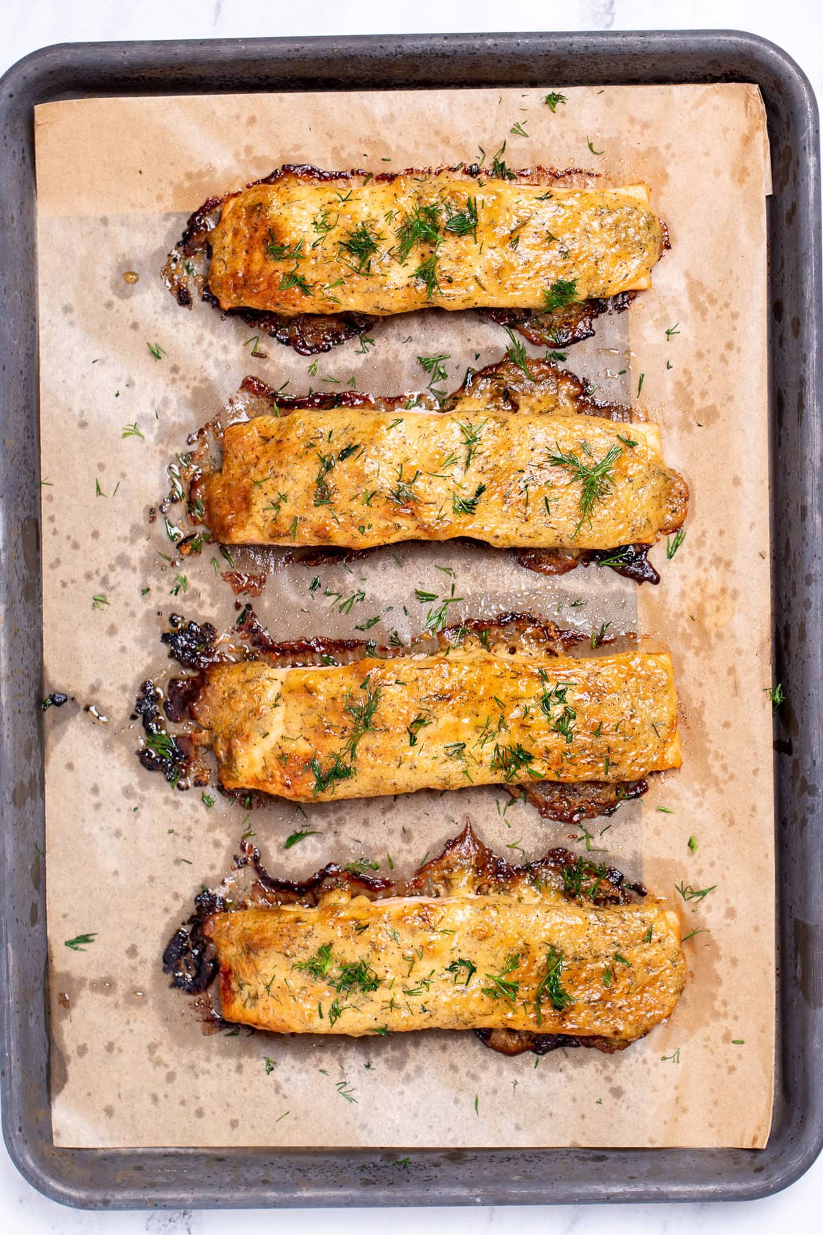 Four baked and broiled salmon fillets topped with honey dijon sauce, on a sheet pan lined with parchment paper, and topped with fresh chopped dill.