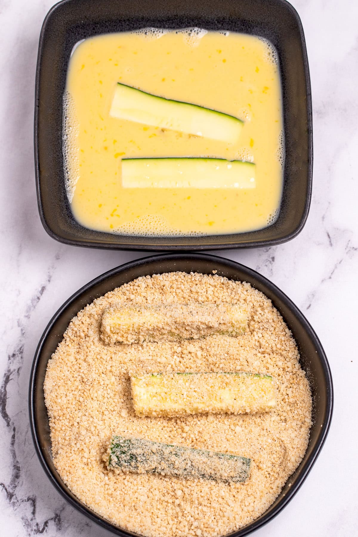 A shallow bowl with zucchini slices being coated in egg, next to another shallow bowl being coated in breadcrumbs and parmesan.